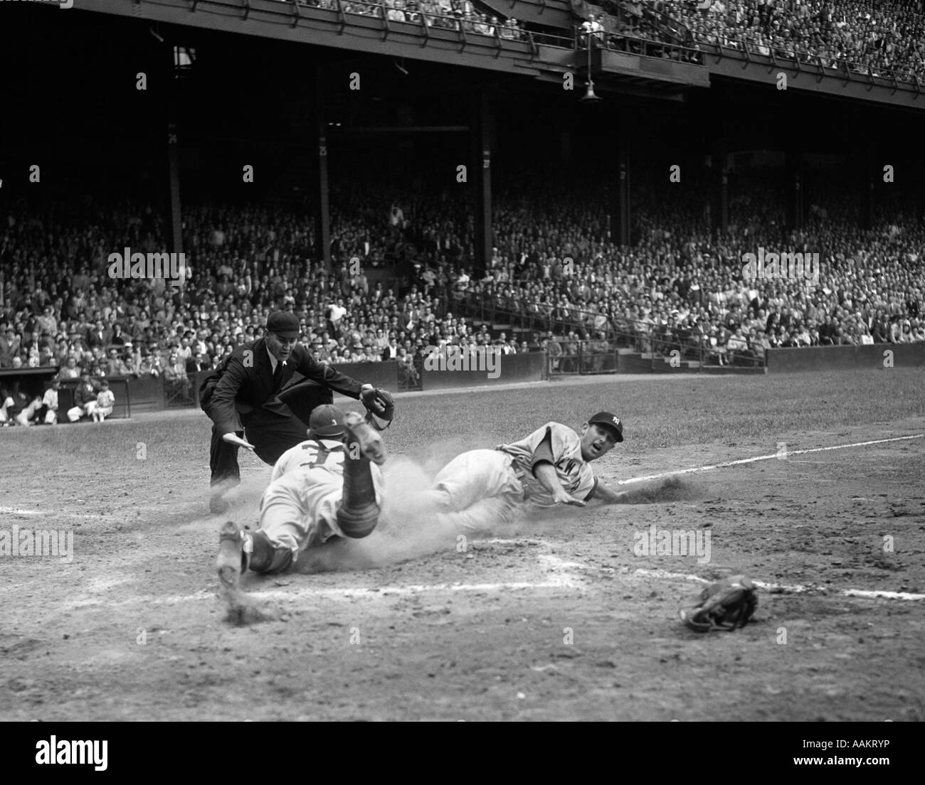 1950s PROFESSIONAL MAJOR LEAGUE BASEBALL GAME RUNNER SLIDING INTO HOME BASE AS UMPIRE SIGNALS SAFE Stock Photo