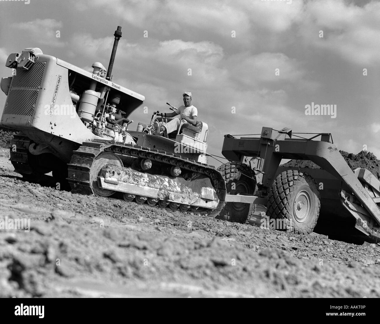 1950s ANGLED WORM'S-EYE VIEW OF MAN PULLING LEVERS ON BULLDOZER HAULING SCRAPER Stock Photo