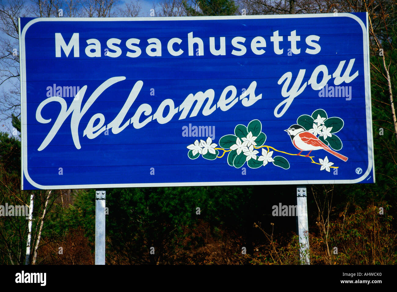 This is a road sign that says Massachusetts welcomes you It is against a blue sky On the sign are the state bird and flowers Stock Photo