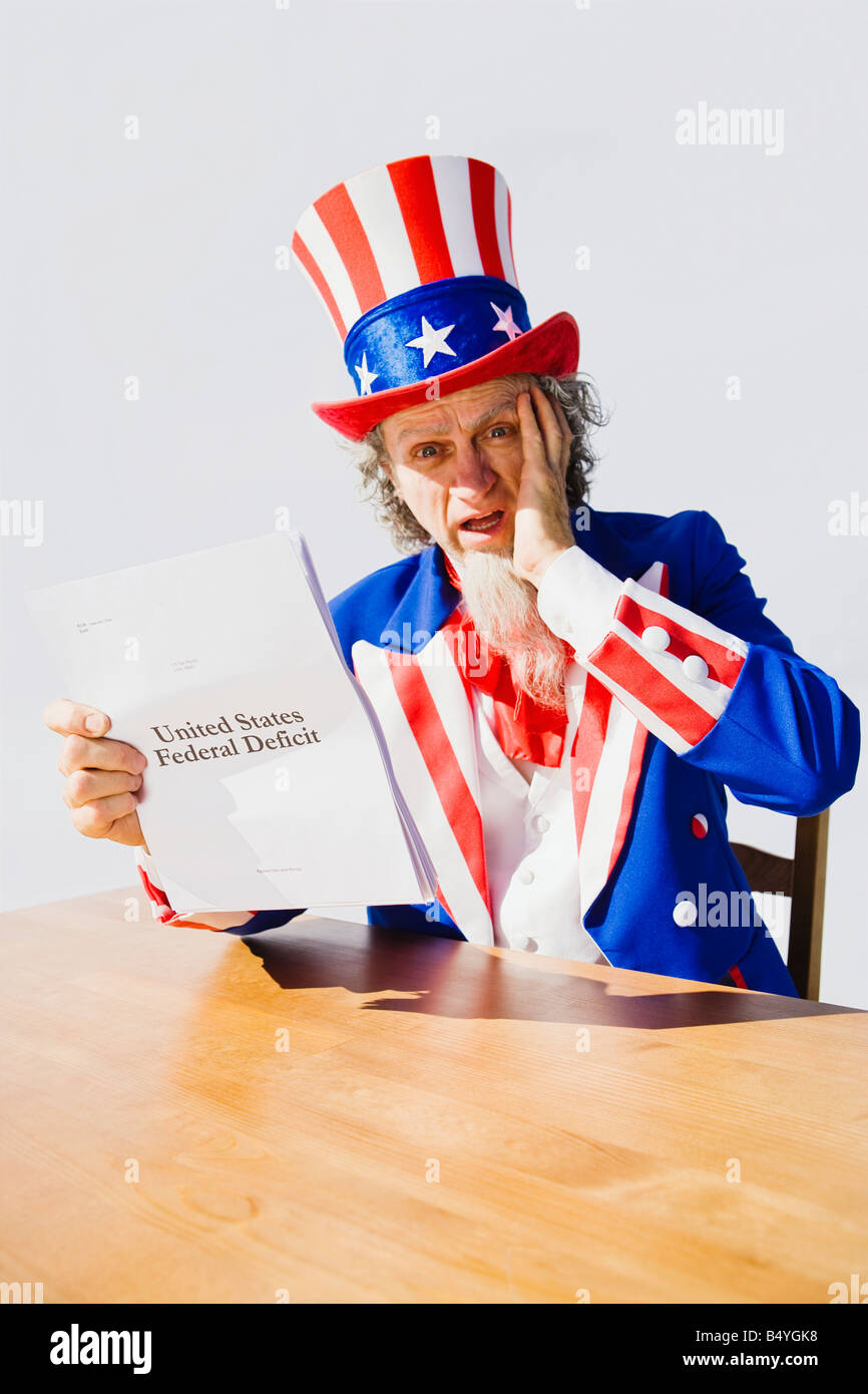 Uncle Sam sitting at a table holding a hand to his head in worry and surprise while looking at the United States Deficit Stock Photo