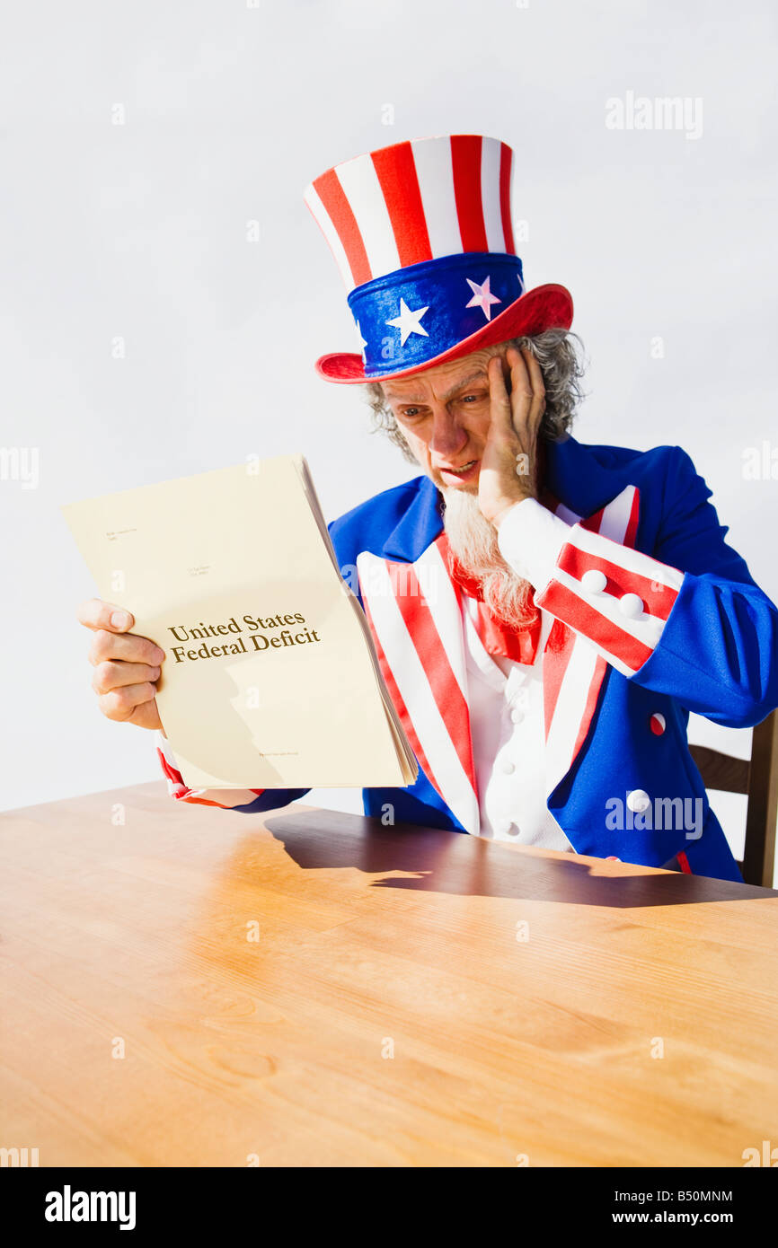 Uncle Sam sitting at a table holding a hand to his head in worry and surprise while looking at the United States Deficit Stock Photo