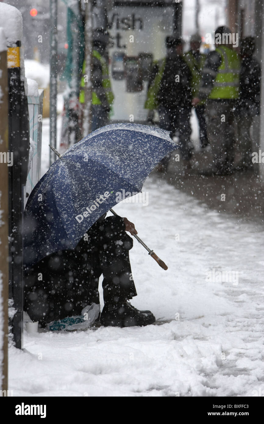 street beggar sitting under deloitte umbrella in the snow on a cold snowy winters day Belfast Northern Ireland Stock Photo