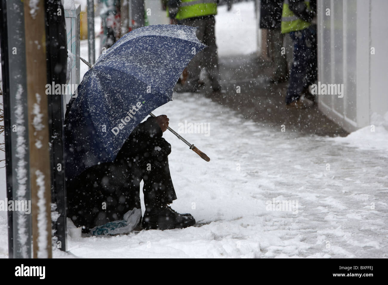 street beggar sitting under deloitte umbrella in the snow on a cold snowy winters day Belfast Northern Ireland Stock Photo