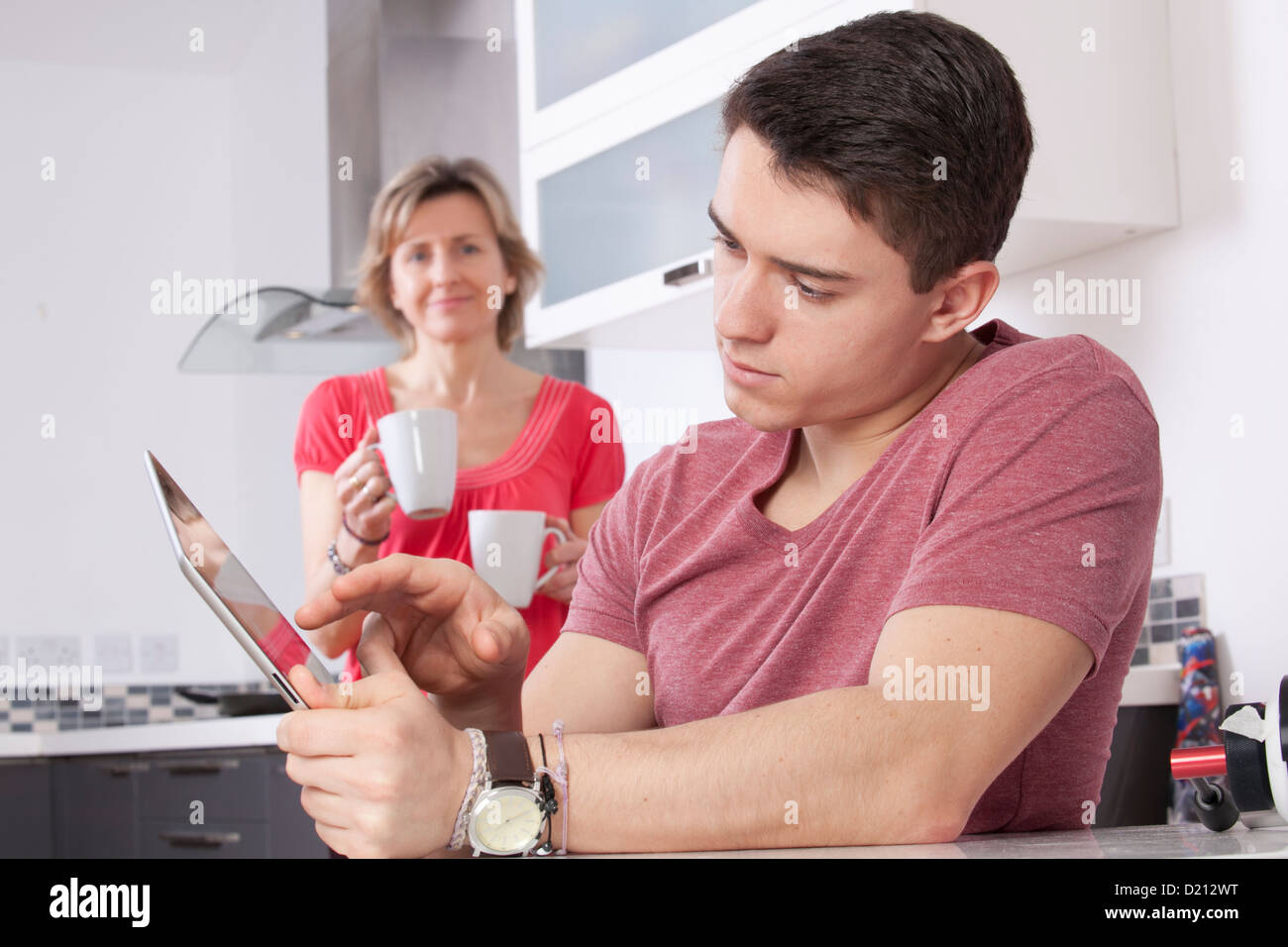 Young man using a digital tablet looking at the screen or monitor. Situated in a modern kitchen a woman holding two mugs smiles. Stock Photo