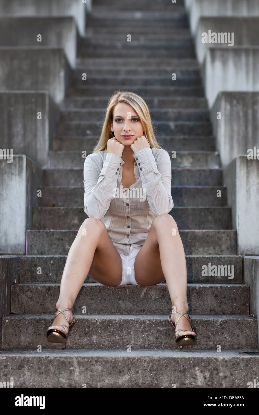 Young woman with long blond hair wearing a grey shirt, white shorts and high heels sitting on stone stairs Stock Photo