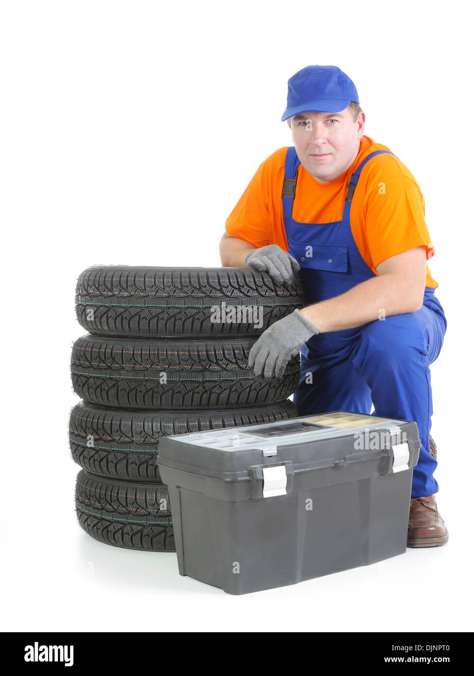 Mechanic wearing blue coveralls and orange t-shirt posing by pile of four new car tires and toolbox shot on white Stock Photo