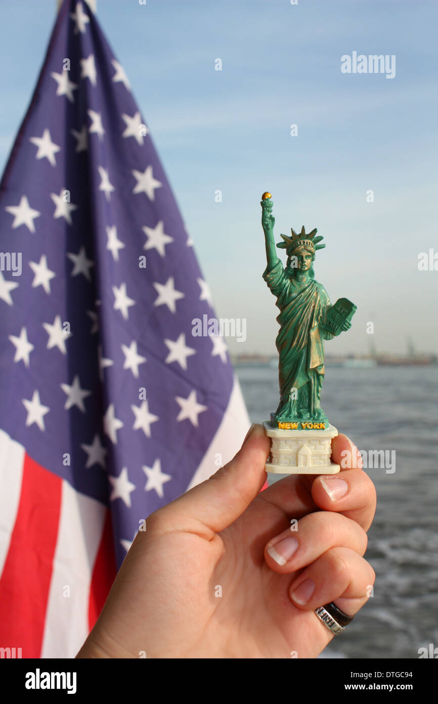 Tourist hand holding a Statue of Liberty souvenir, overlooking the water, red & blue American flag in the background. Stock Photo