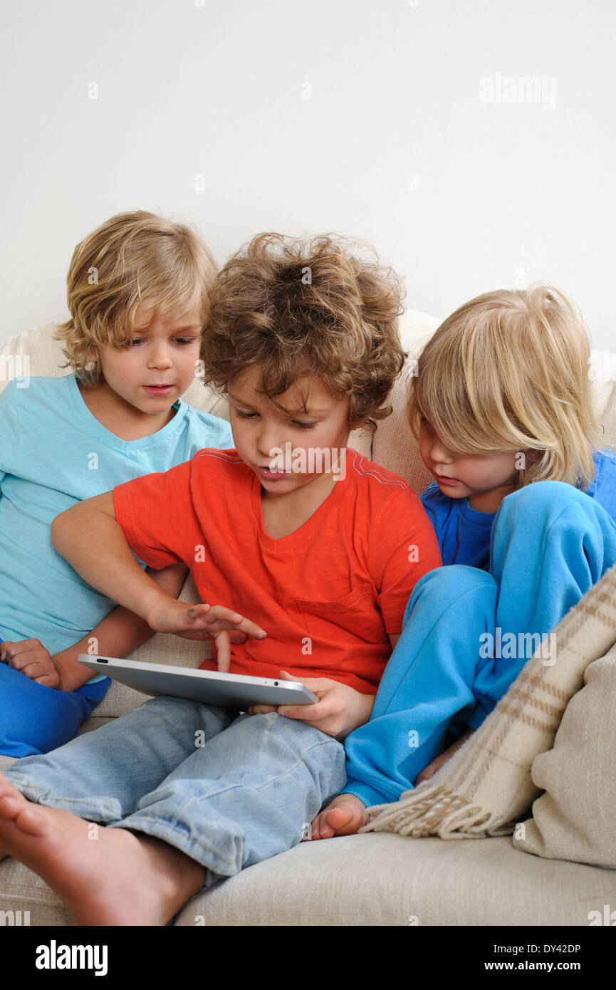 An 8-9 year old is playing a game on a touch screen tablet. His younger brothers are watching every move intently Stock Photo