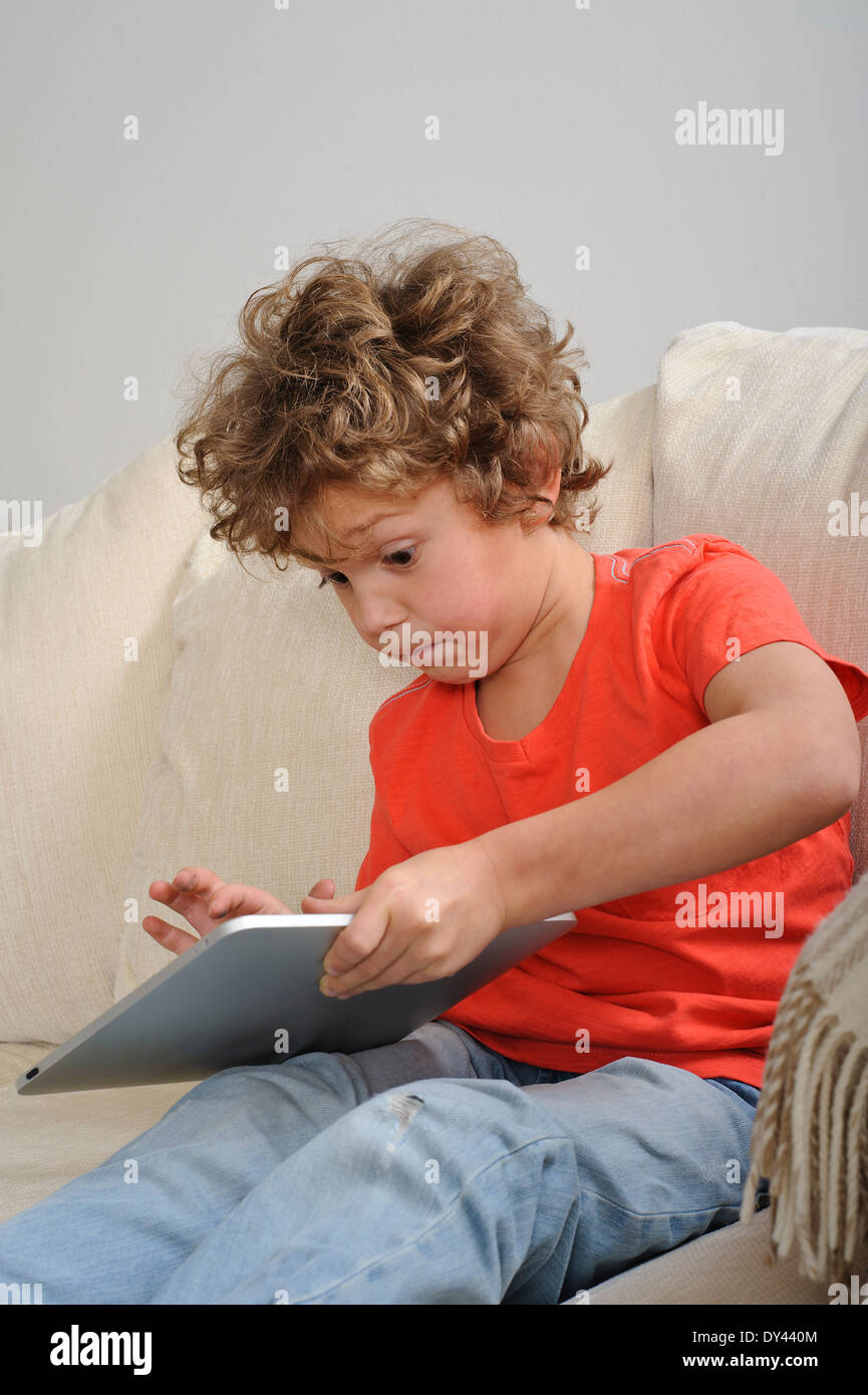 An 8-9 year old boy plays games on a touch screen tablet. He's sitting on a sofa Stock Photo