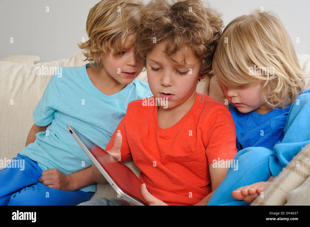 An 8-9 year old is playing a game on a touch screen tablet. His younger brothers are watching every move intently Stock Photo