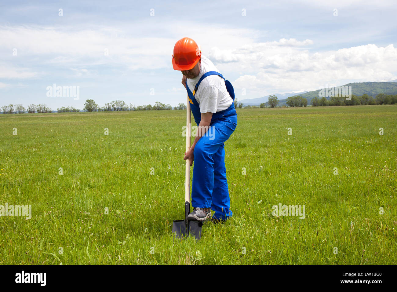 Worker in coveralls is digging with a shovel, in a green meadow. He is starting a new construction project. The image was shoote Stock Photo