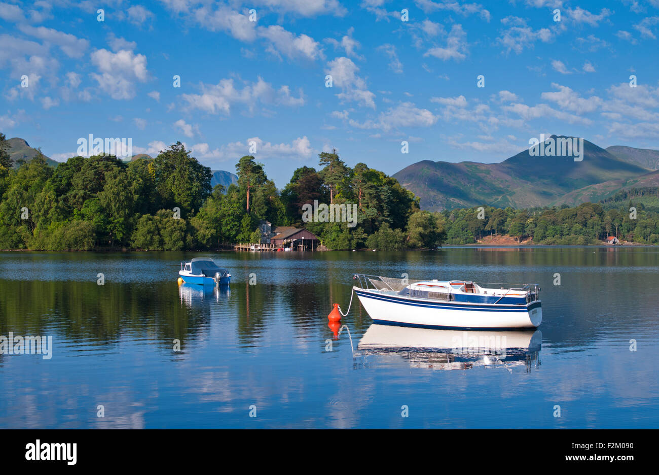 View over small boats moored on Derwentwater to wooded Derwent Island, fells rising behind, calm sunny morning, Cumbria UK Stock Photo