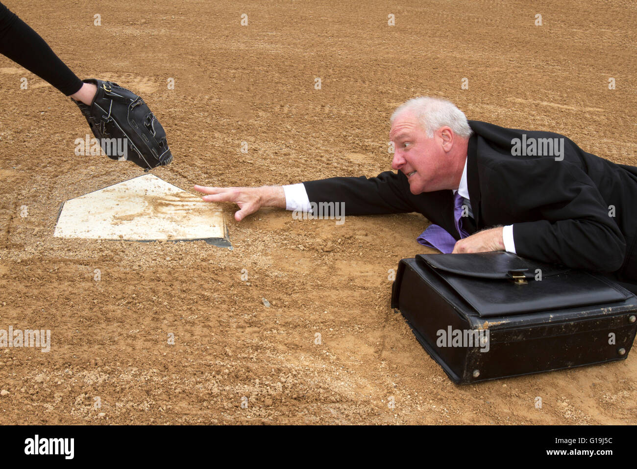 Businessman in suit and tie with a briefcase trying to slide into home plate with a glove waiting to tag him out. Stock Photo