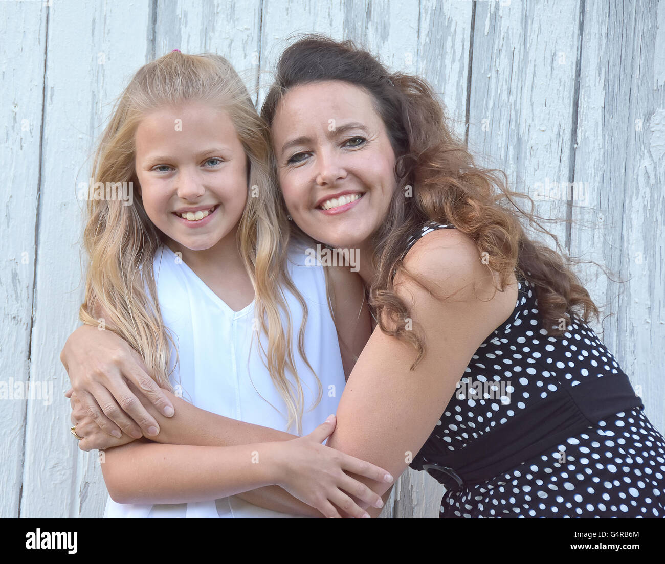 Smiling Caucasian mother hugging little blond daughter by old whitewashed barn. Stock Photo