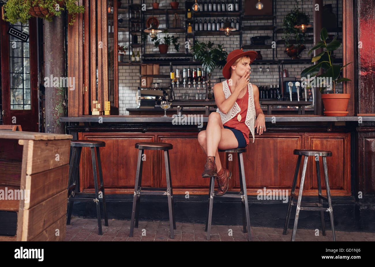 Young woman with hat smoking in a bar. Holding cigarette and looking away. Stock Photo