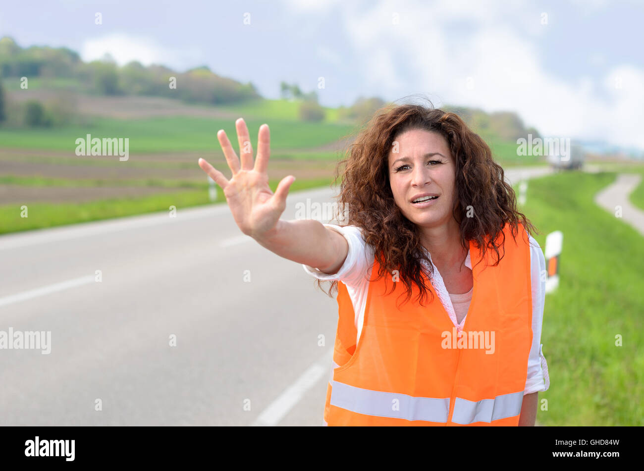 Frantic young woman in a high visibility jacket standing at the side of a rural road trying to stop traffic by raising her palm  Stock Photo