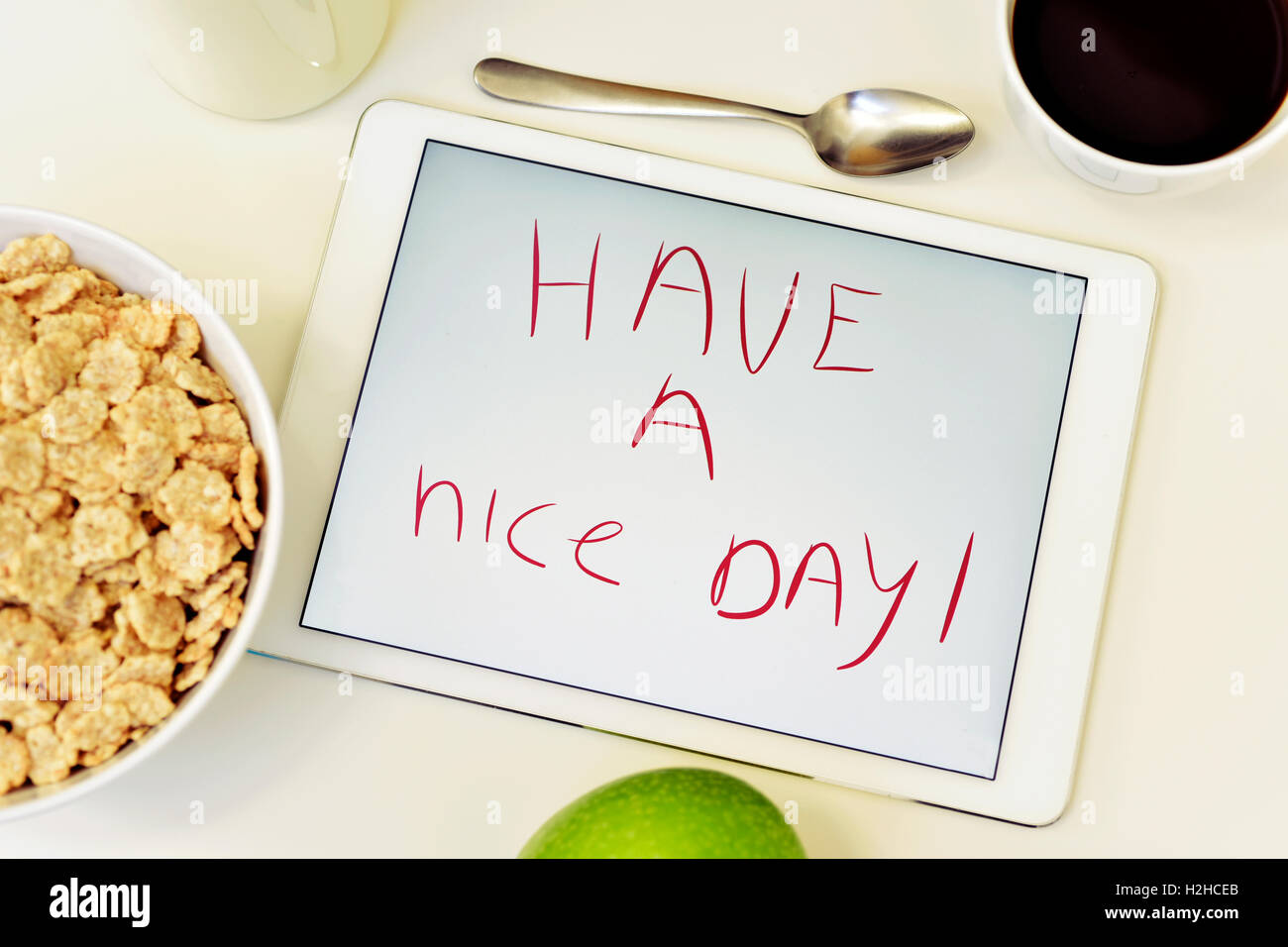 the text have a nice day in a tablet, placed on a white table set for breakfast with a green apple, a cup of coffee and a bowl w Stock Photo