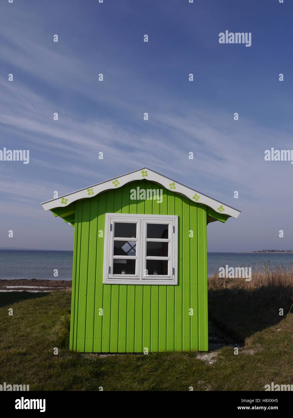 A cute lime-green beach hut with white roof and window frame against a background of blue sea and blue sky. Stock Photo