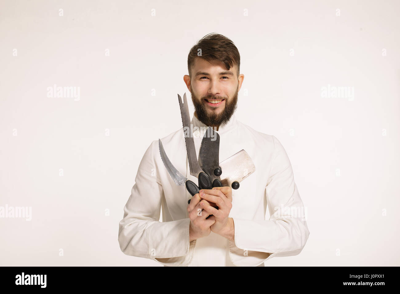 Studio shot of a happy bearded young chef holding sharp knives over white background. Chef with knife. Handsome smiling cheef holding many sharp knive Stock Photo