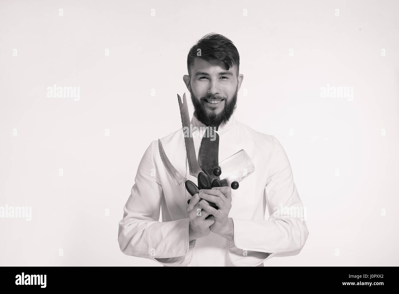 Studio shot of a happy bearded young chef holding sharp knives over white background. Chef with knife. Handsome smiling cheef holding many sharp knive Stock Photo