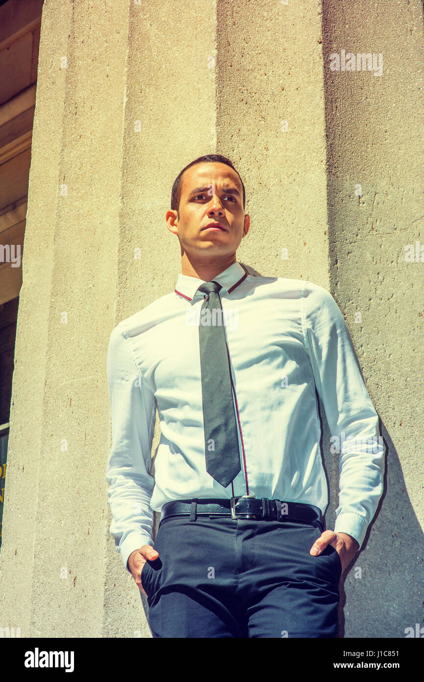 Young American Businessman wearing white shirt, black tie, standing against column outside office building in New York, under strong sun, frowned, thi Stock Photo