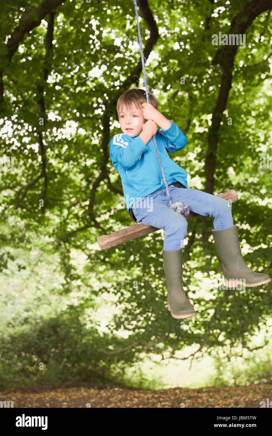 Boy in wellies swinging on tree rope swing Stock Photo