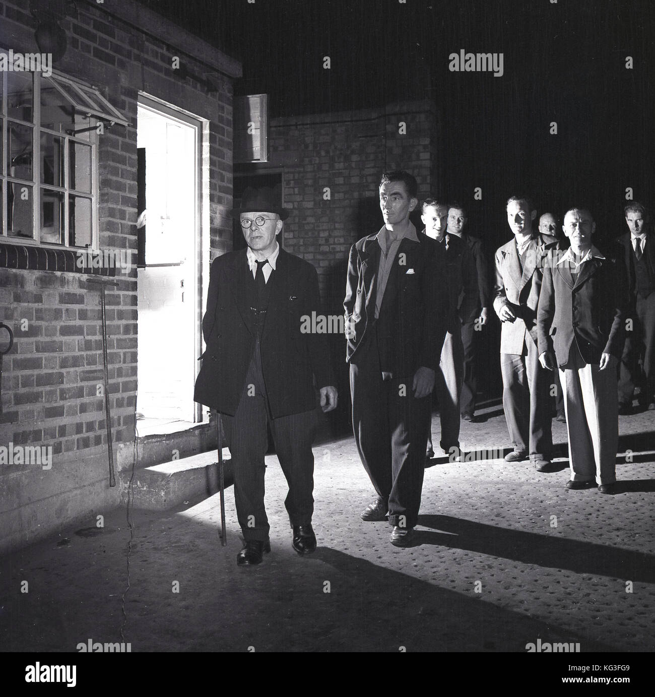 1950s, historical, England, 'social class'....group of men mingling outside a working men's club create an uneasy, menacing atmosphere for an elderly city gentleman going by them, who they consider a 'toff', with his top hat, blazer, pinstripe trousers and walking stick. Stock Photo