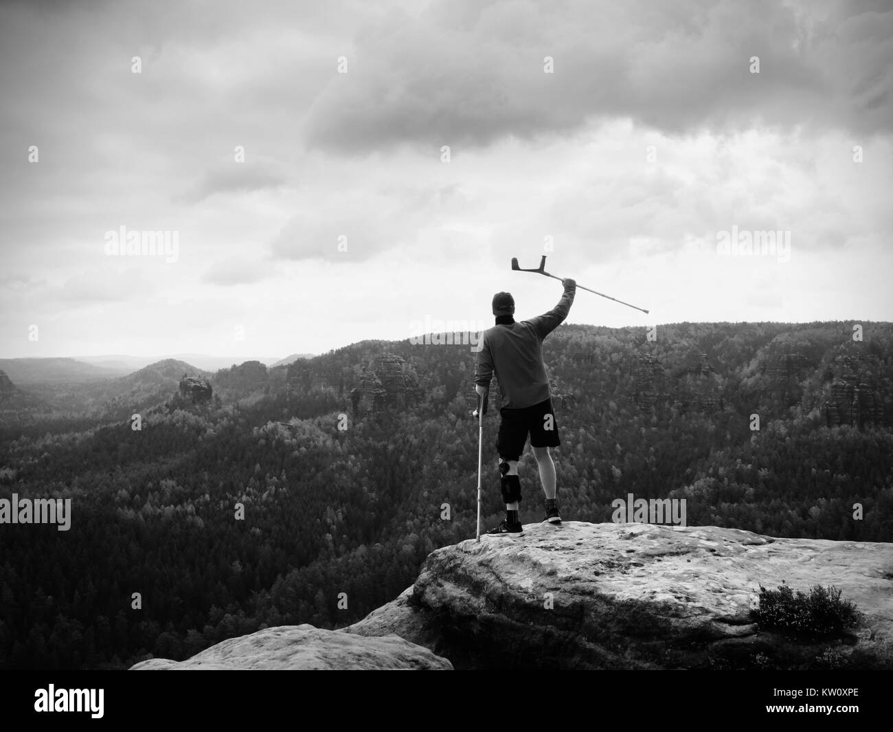 Man with forearm crutch. Hiker finaly achieved mountain peak. Tourist with broken leg in immobilizer hold medicine  poles. Stock Photo