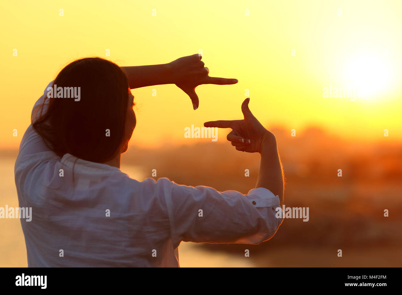 Back view backlight portrait of a woman framing with fingers at sunset Stock Photo