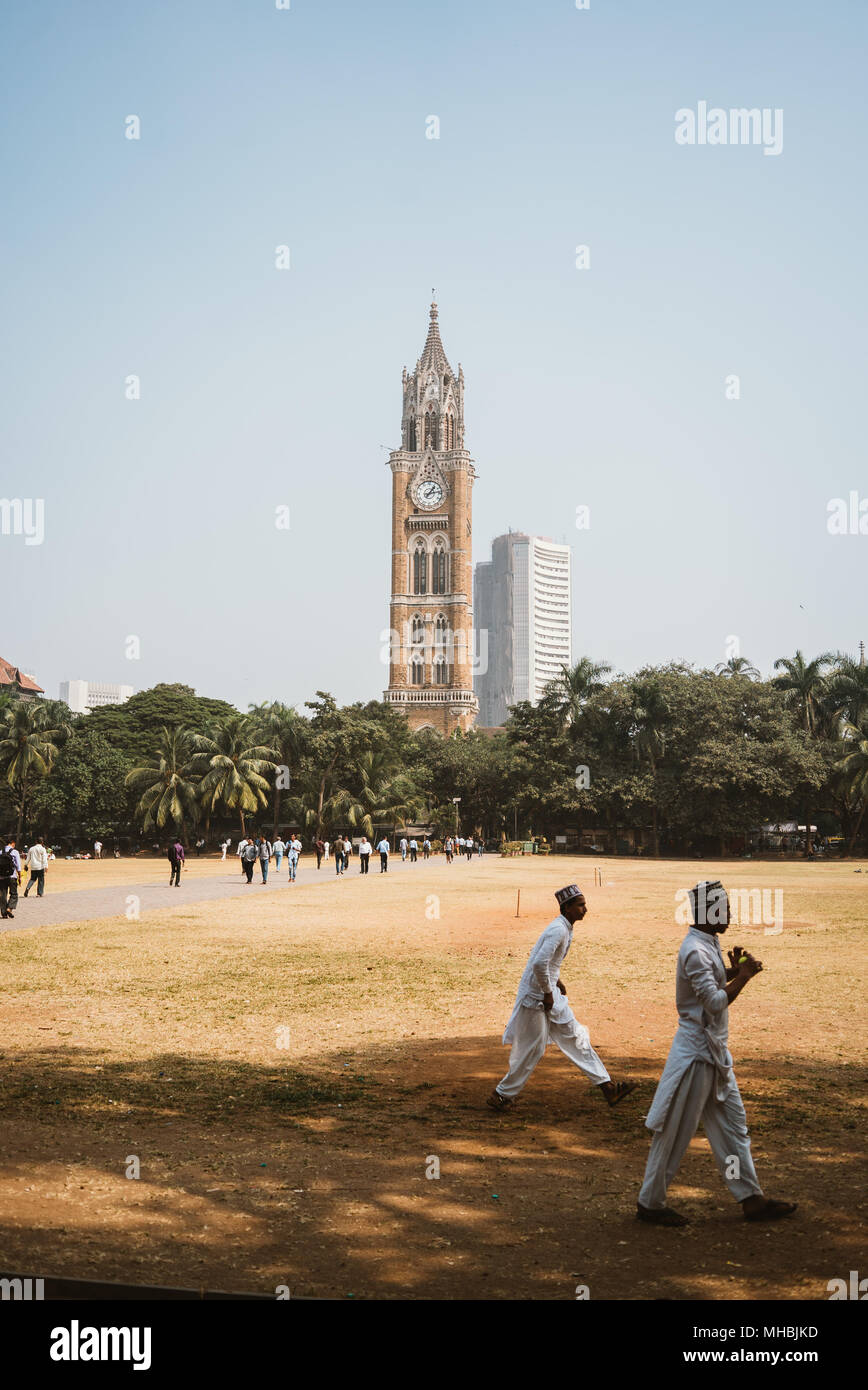 Indian boys playing cricket in a dry field with a gothic clock tower in the distance Mumbai India Stock Photo