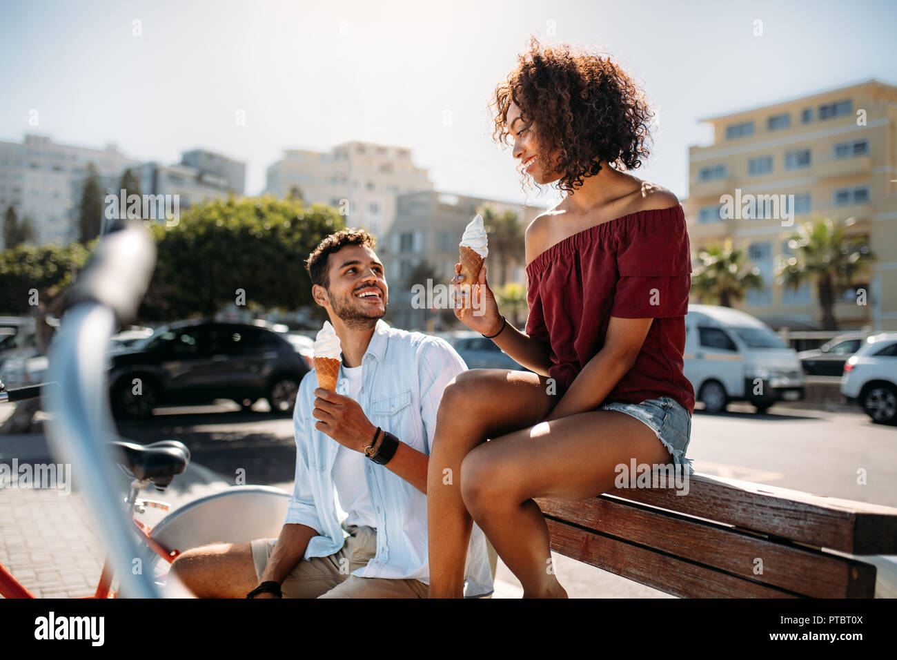 Romantic couple sitting on a bench in street eating cone ice cream looking at each other. Young man and woman having fun moving around the city eating Stock Photo