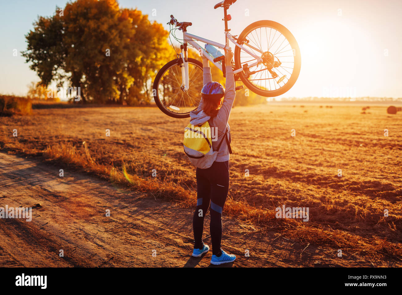 Young bicyclist raising her bicycle in autumn field at sunset. Happy ...