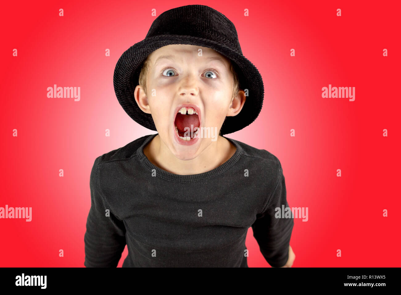 closeup of young boy in black shirt and black hat with heavy screaming expression and eyes wide open with crazy look on red background Stock Photo