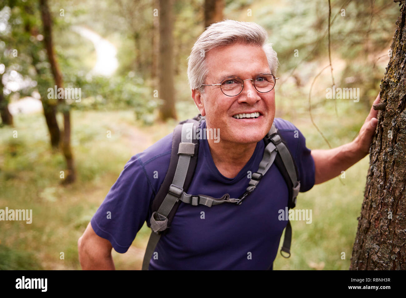 Middle aged Caucasian man taking a break during a hike, leaning on a tree in a forest, waist up, close up Stock Photo