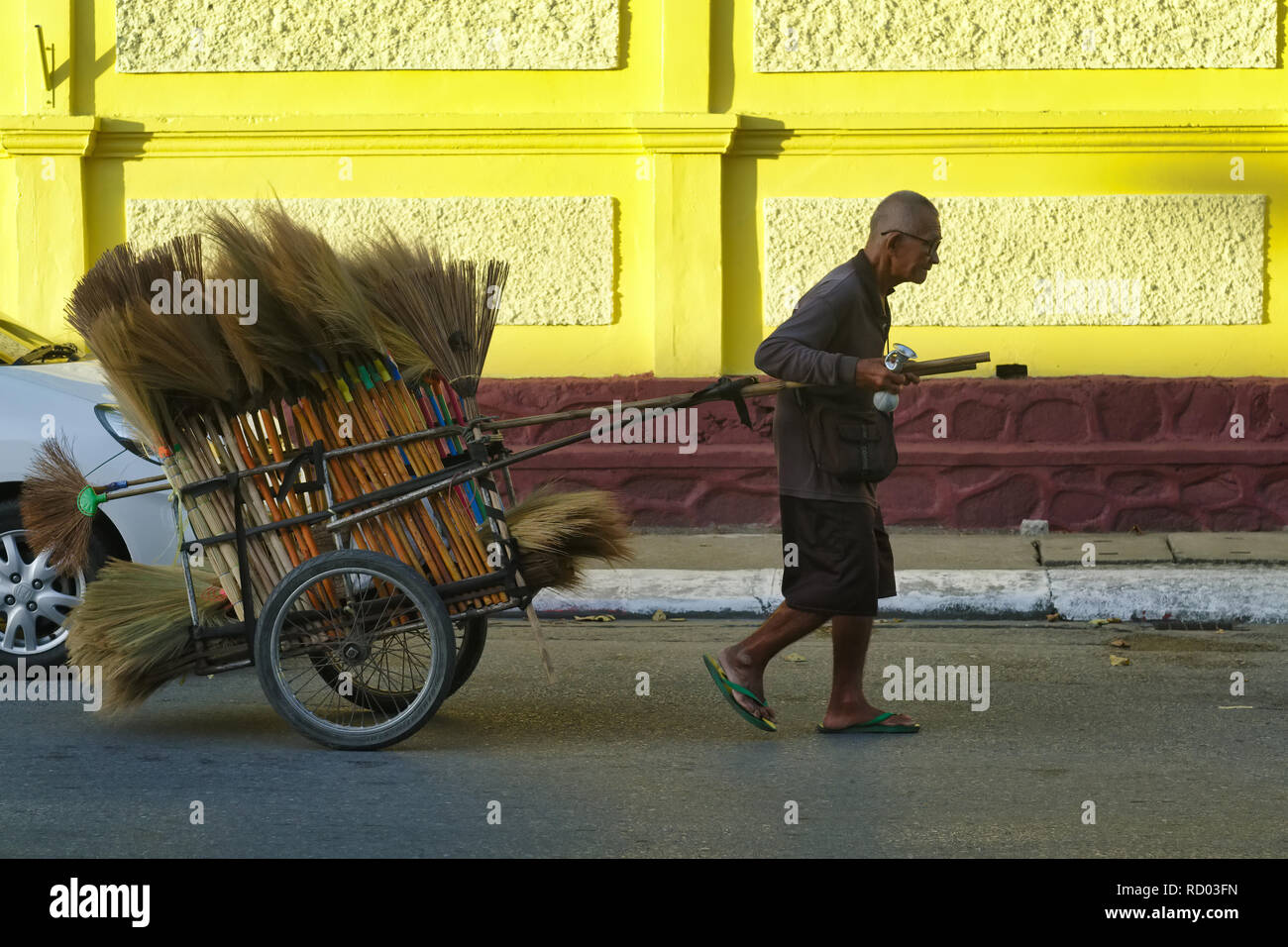 A elderly man in Phuket Town, Phuket, Thailand. pulls a cart full of handmade brooms past the yellow wall of Wat Mongkon Nimit Stock Photo