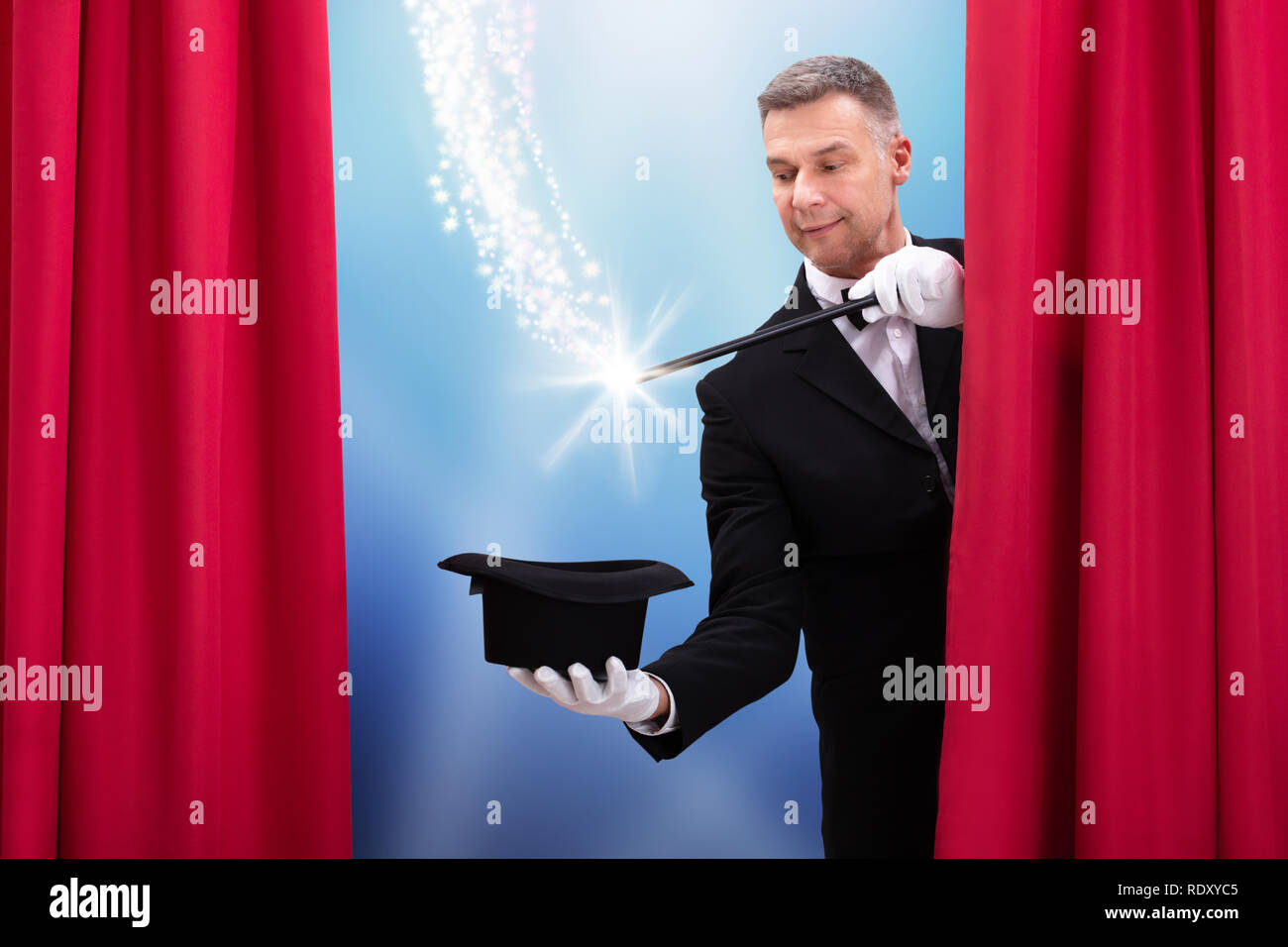 Magician Doing Magic Trick With Illuminated Wand And Hat Behind Curtain Stock Photo