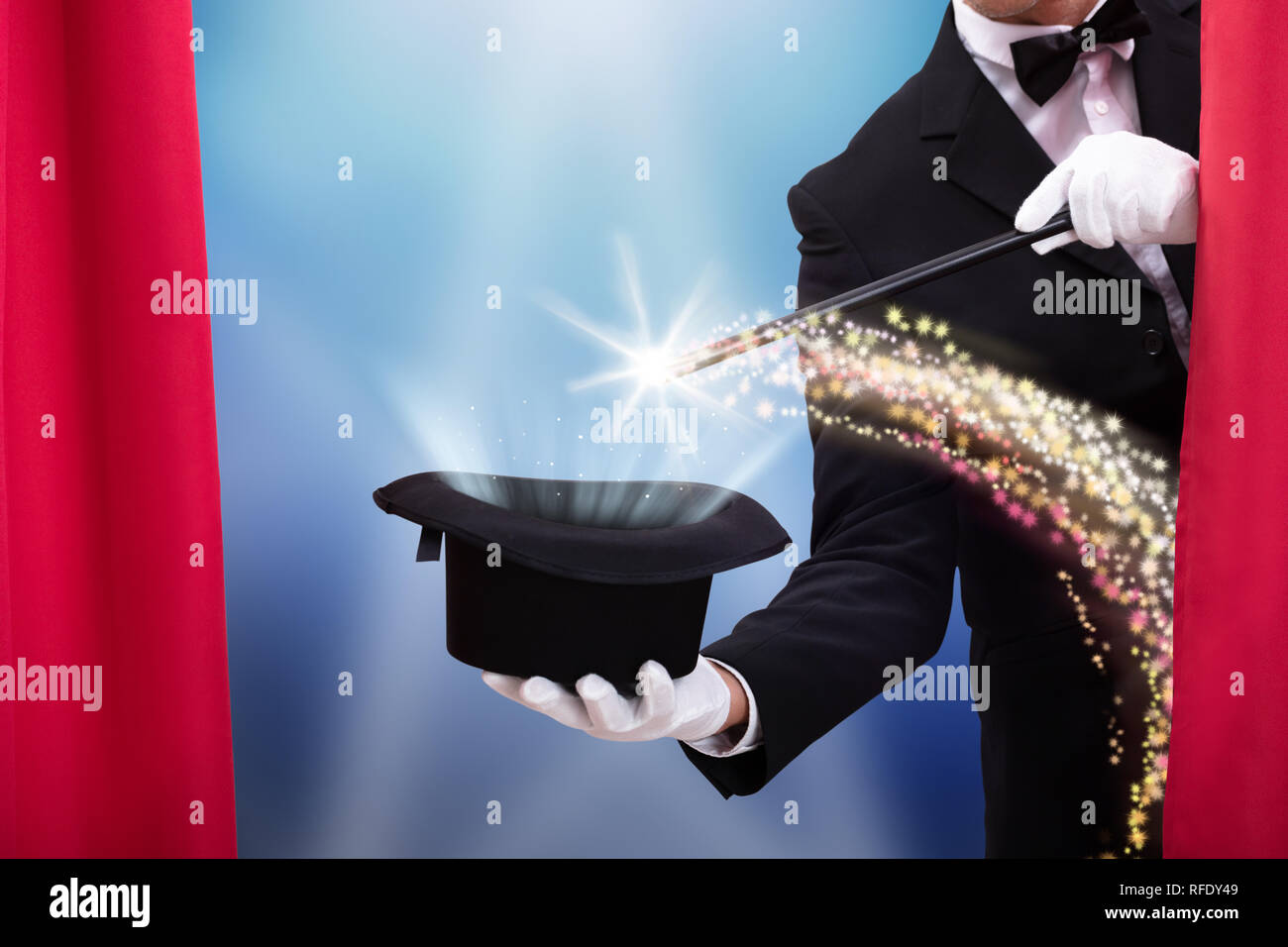 Magician Doing Magic Trick With Illuminated Wand And Hat Behind Curtain Stock Photo