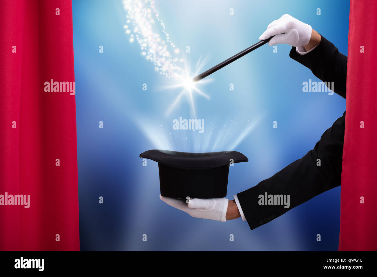 Magician Doing Magic Trick With Illuminated Wand And Hat Behind Curtain Stock Photo