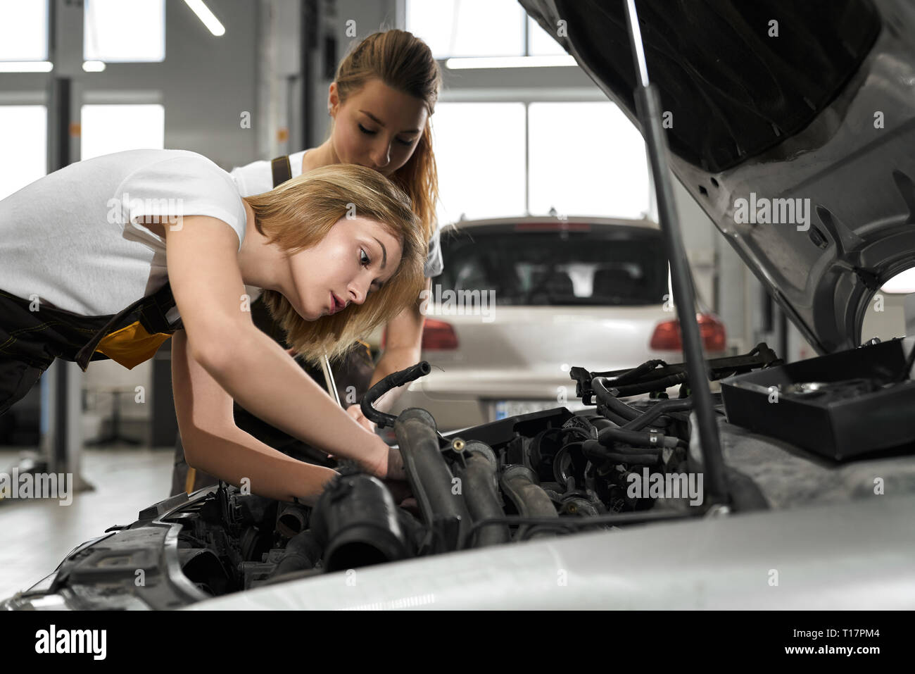 Two mechanics repairing and working maintenance of car in autoservise. Young beautiful blonde girls wearing in coveralls and white t shirts looking at car with opened hood. Stock Photo