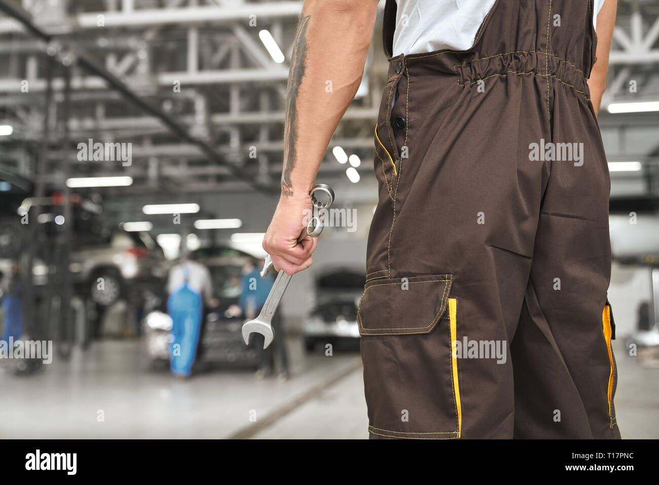 Back view of mechanic standing in autoservice, holding wrench, tool for fixing automobiles. Repairman wearing in brown coveralls, uniform. Stock Photo