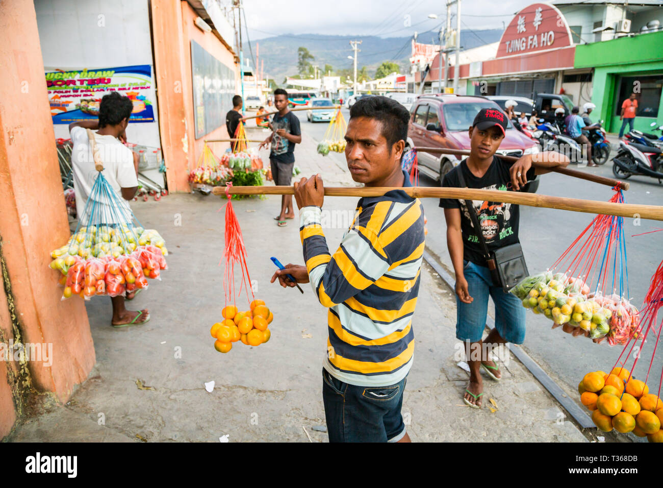 Timorese men, street vending fruits and vegetables hanging on ropes from neck yokes made of sticks on a busy market street of capital Dili, East Timor. Stock Photo