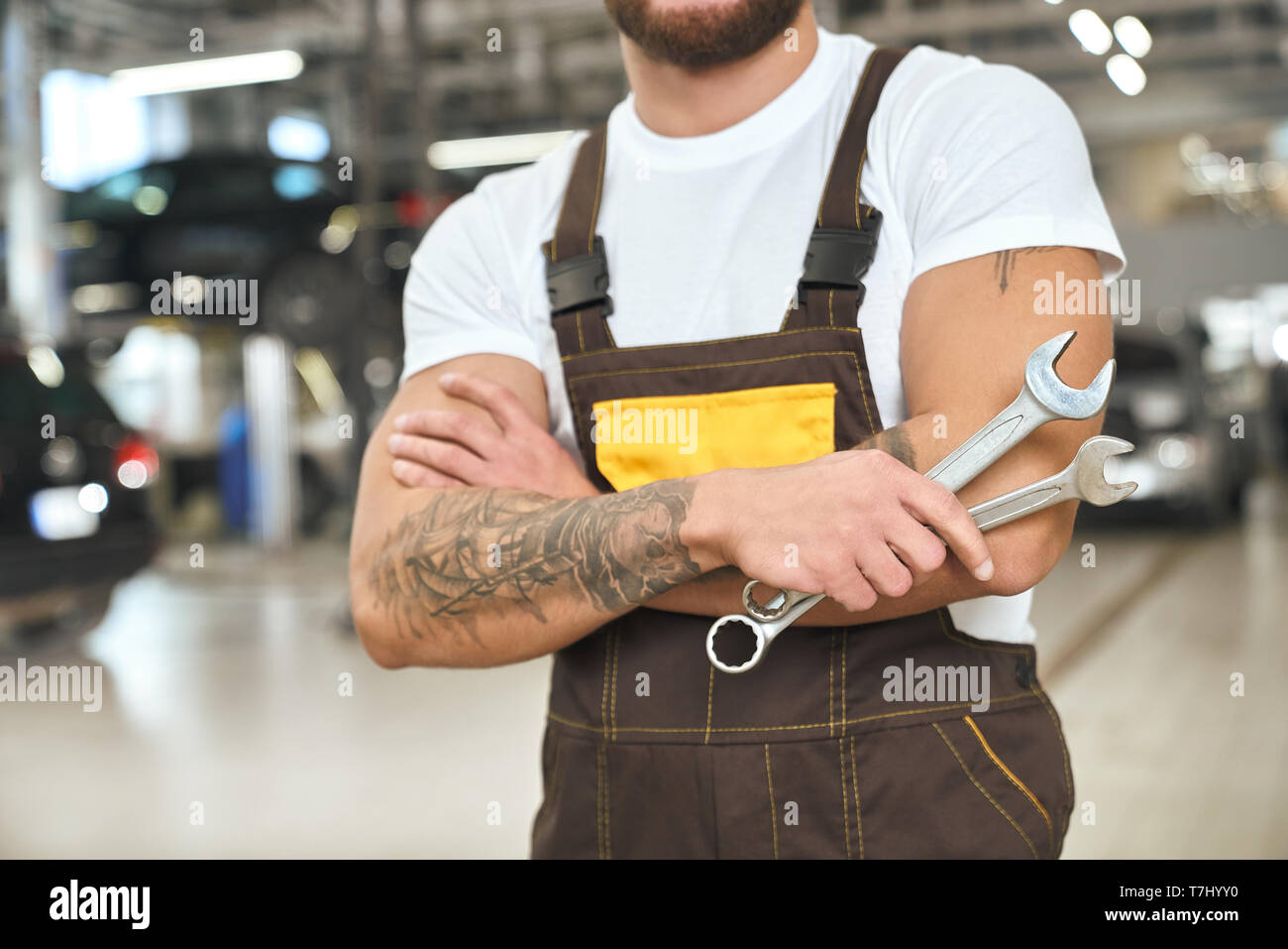 Unrecognizable muscular mechanic with tattoed hands, wearing in coveralls and white t shirt. Brutal man holding wrenches, working in autoservice. Stock Photo