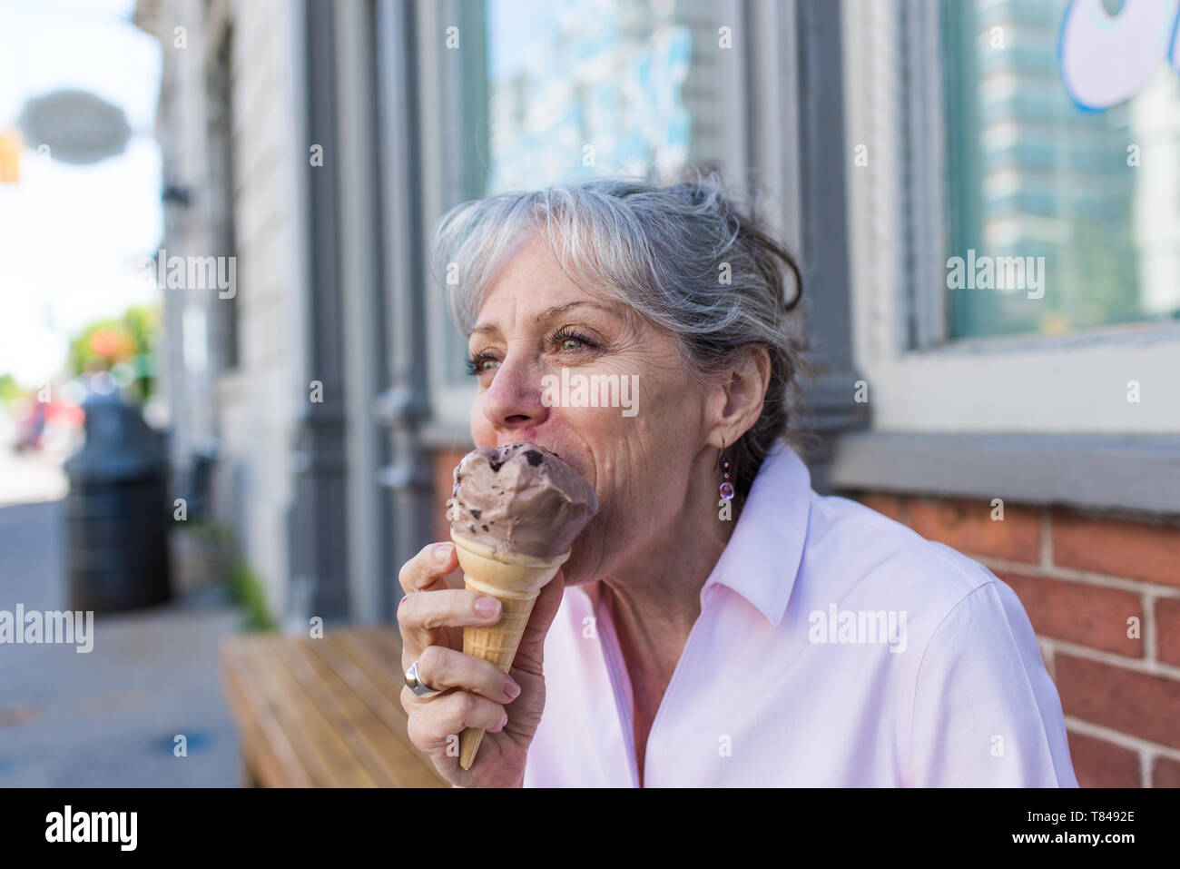 Senior woman sitting on sidewalk eating chocolate ice cream cone Stock Photo