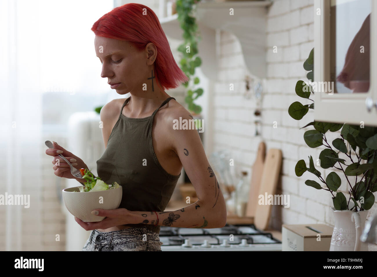 Skinny red-haired woman with tattoos eating vegetable salad Stock Photo