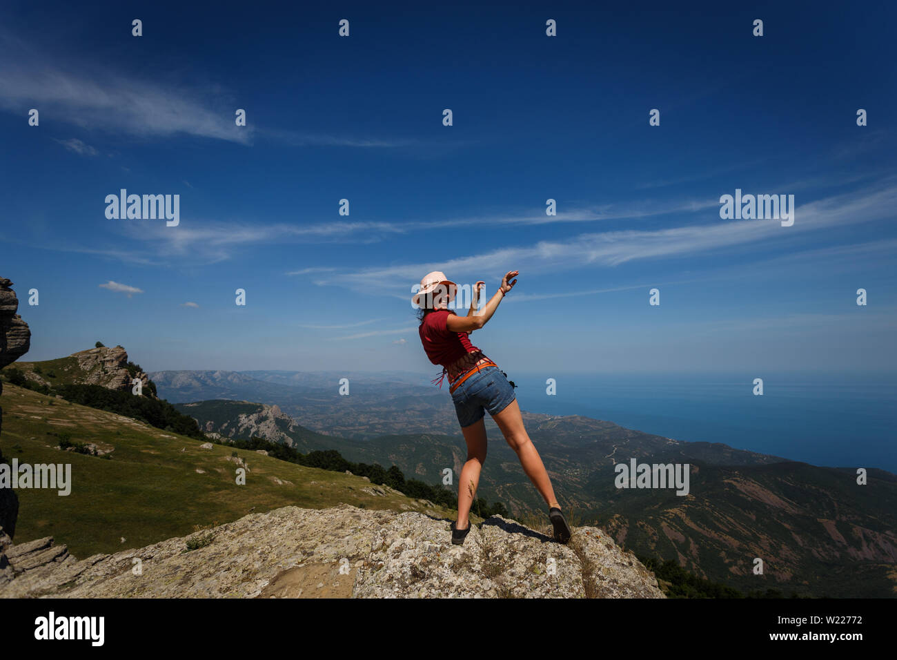 Happy woman in short shorts and wearing a hat is dancing on the top of a mountain. The concept of freedom, recreation, travel. Good luck and success. Stock Photo