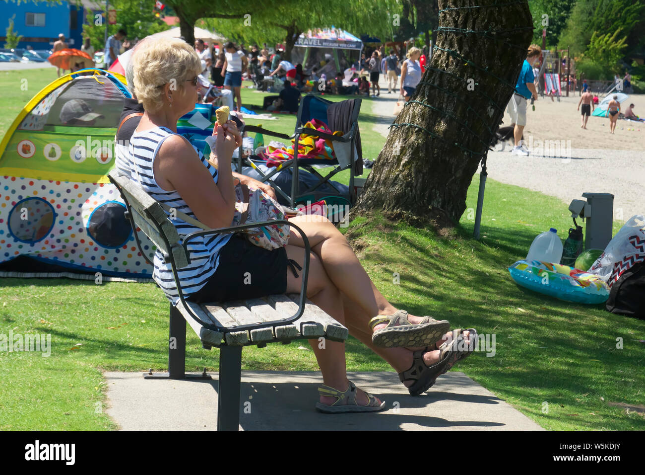 Blonde woman sitting on a bench enjoying an ice cream cone with many people in the background at Harrison Hot Springs July 2019. Stock Photo