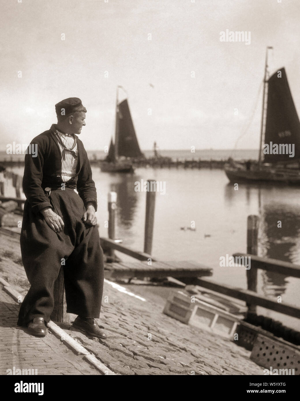 1920s MAN FISHERMAN IN TRADITIONAL CLOTHES SAIL BOATS IN BACKGROUND VOLENDAM HARBOR HOLLAND  - r3740 HAR001 HARS TRANSPORTATION B&W HARBOR SKILL OCCUPATION SKILLS TROUSERS SAILBOATS TOURIST IN ATTRACTION OCCUPATIONS HOLLAND CONCEPTUAL POPULAR STYLISH FISHING BOAT MID-ADULT MID-ADULT MAN BLACK AND WHITE CAUCASIAN ETHNICITY HAR001 NETHERLANDS OLD FASHIONED Stock Photo