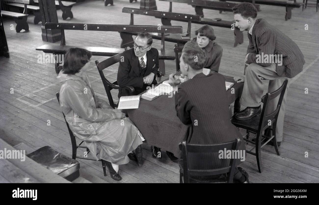 Années 1950, historique, à l'intérieur d'une salle d'église, une fille dans un rainsmac assis autour d'une table avec ses parents, frère et un enseignant, avec des livres dehors, discutant peut-être des chapitres de la bible, Witney, Oxford, Angleterre, Royaume-Uni. Banque D'Images