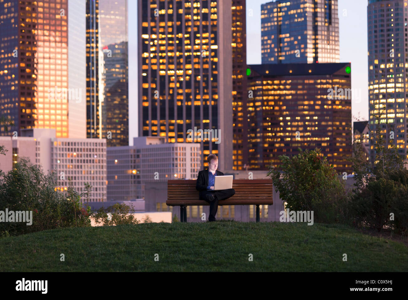 Voce maschile seduto su una panchina nel parco lavorando sul computer portatile con Los Angeles skyline della città sullo sfondo al tramonto tramonto Foto Stock