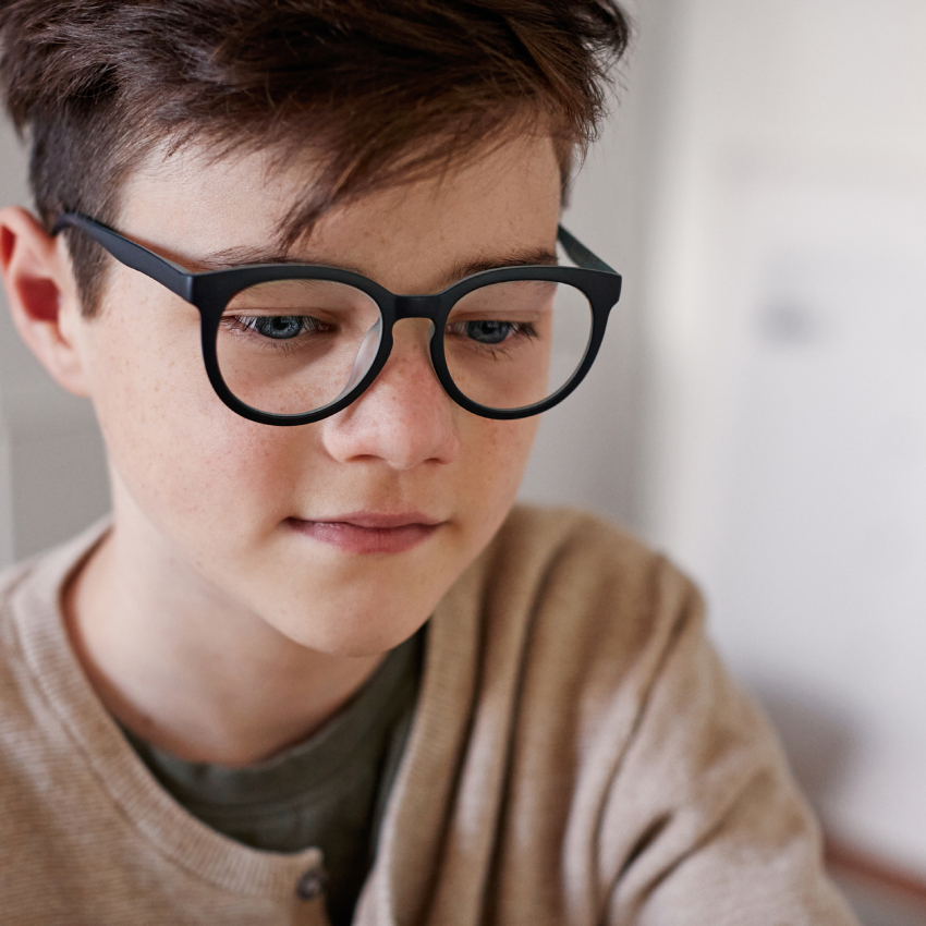 Boy wearing black, round glasses and a beige cardigan.